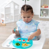 A toddler wearing a pastel bib in a high chair reaches for snacks from a Bumkins Silicone Grip Dish: Blue Jelly while holding blue utensils.