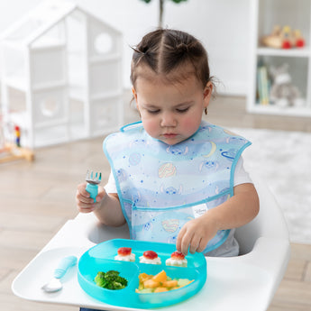 A toddler wearing a pastel bib in a high chair reaches for snacks from a Bumkins Silicone Grip Dish: Blue Jelly while holding blue utensils.