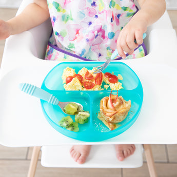 A child in a high chair enjoys eggs, tomatoes, kiwi, and pancakes from a Bumkins Silicone Grip Dish: Blue Jelly.