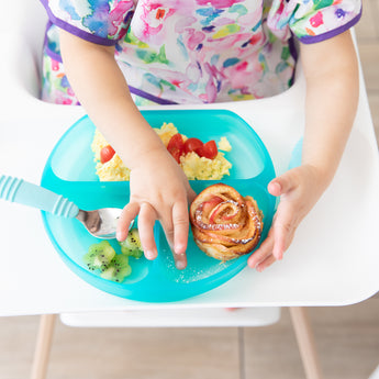 A child in a high chair wearing a colorful bib enjoys kiwi, eggs, tomatoes & a pastry from a Bumkins Silicone Grip Dish: Blue Jelly.