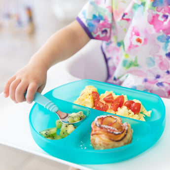 A child eats kiwi, eggs with tomatoes, and a pastry from a Bumkins Silicone Grip Dish: Blue Jelly while wearing a colorful bib.