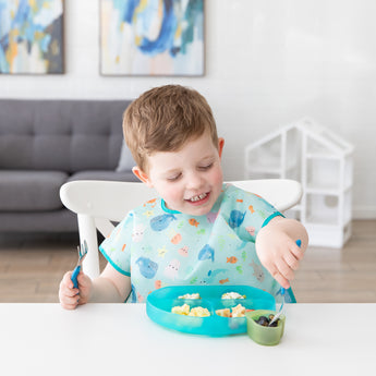 A child eagerly eats from a Bumkins Silicone Grip Dish: Blue Jelly on a white table in a bright room.