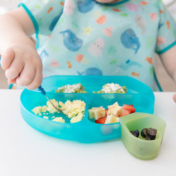 A child uses a Bumkins Silicone Grip Dish: Blue Jelly for eggs and avocado, with berries in a green cup, wearing a sea-themed bib.