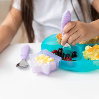 A child enjoys breakfast from a Bumkins Silicone Grip Dish: Blue Jelly, using a lavender fork, with yogurt and pineapple in a star bowl.