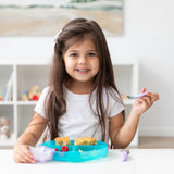 A young girl with long brown hair smiles while eating from a Bumkins Silicone Grip Dish: Blue Jelly at a white table.