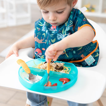 A boy in a jungle themed bib sits in a high chair, enjoying a meal from a Bumkins Silicone Grip Dish: Blue Jelly with orange utensils.