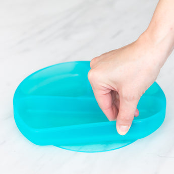 A hand holds a Bumkins Silicone Grip Dish: Blue Jelly, demonstrating the suction cup bottom on a marble surface.