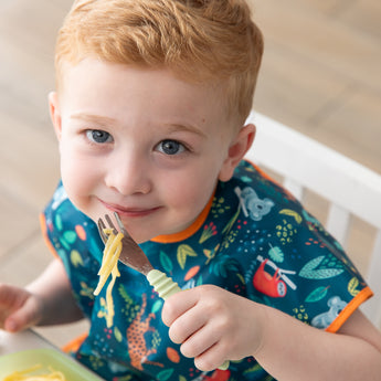 A child in a dark blue bib smiles at the camera, holding a Bumkins Spoon + Fork: Sage with spaghetti.