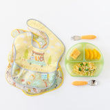 Colorful bibs sit next to Bumkins Silicone Grip Dish: Green Jelly, holding pasta, cornbread, and oranges, and utensils on a white background.