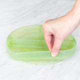 A hand holds a Bumkins Silicone Grip Dish: Green Jelly, demonstrating the suction cup bottom on a marble surface.