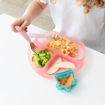 A child enjoys a meal from a Bumkins Silicone Grip Dish: Pink Jelly filled with pasta, broccoli, and apples, with peanut butter in a star bowl.