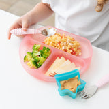 A child at a white table enjoys a meal from a Bumkins Silicone Grip Dish: Pink Jelly. A nearby blue star-shaped dish holds peanut butter.