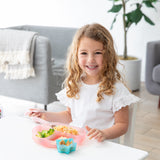 A curly-haired girl smiles while eating from a Bumkins Silicone Grip Dish: Pink Jelly at a white table, with a plant and sofa behind her.