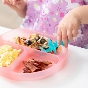 Close-up of a child eating waffles and berries with a blue fork from a Bumkins Silicone Grip Dish: Pink Jelly at a white table.