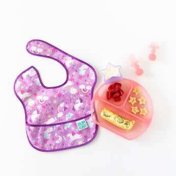 A bib next to Bumkins Silicone Grip Dish: Pink Jelly with berries, quiche, and banana, a yogurt cup, and utensils on a white background.