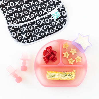 Bumkins Silicone Grip Dish: Pink Jelly with quiche and fruit sits next to a bib, utensils, and a star cup of yogurt on a white background.