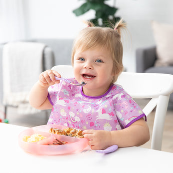 A toddler with pigtails is smiling at a white table, wearing a purple bib and enjoying a meal from a Bumkins Silicone Grip Dish: Pink Jelly.