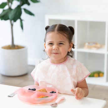A girl in a pink bib with pigtails smiles; a dish from Bumkins Grip Dish Deluxe Infant 5-Piece Mealtime Set: Pink Jelly sits in front of her.