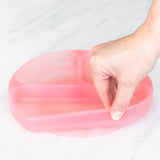 A hand holds a Bumkins Silicone Grip Dish: Pink Jelly, demonstrating the suction cup bottom on a marble surface.