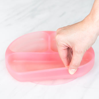 A hand holds a Bumkins Silicone Grip Dish: Pink Jelly, demonstrating the suction cup bottom on a marble surface.