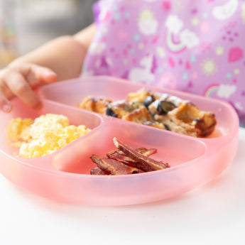 Close-up of a child with a Bumkins Silicone Grip Dish: Pink Jelly filled with scrambled eggs, bacon, and blueberry-topped waffles.