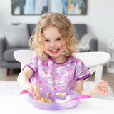 A smiling curly-haired child enjoys food from a Bumkins Silicone Grip Dish: Purple Jelly at a white table, wearing a pink unicorn-themed bib.