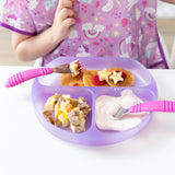 A child in a colorful unicorn bib sits at a white table with a Bumkins Silicone Grip Dish: Purple Jelly filled with pancakes and yogurt.