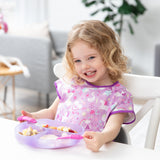 A smiling child in a pink bib sits eagerly at a white table with a Bumkins Silicone Grip Dish: Purple Jelly, ready for mealtime.
