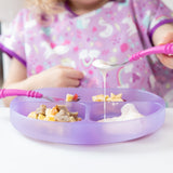 A child in a purple bib enjoys yogurt from a Bumkins Silicone Grip Dish: Purple Jelly using pink utensils.