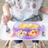 A child in a high chair enjoys scrambled eggs, bananas, and fruit salad from a Bumkins Silicone Grip Dish: Purple Jelly using a spoon.