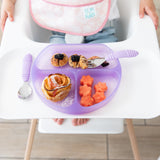A child in a high chair has a Bumkins Silicone Grip Dish: Purple Jelly with cinnamon rolls, melon flowers, berries, and purple utensils.