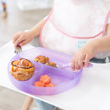 A child with a bib sits at a highchair, holding a fork and Bumkins Silicone Grip Dish: Purple Jelly with sweet rolls and fruit.