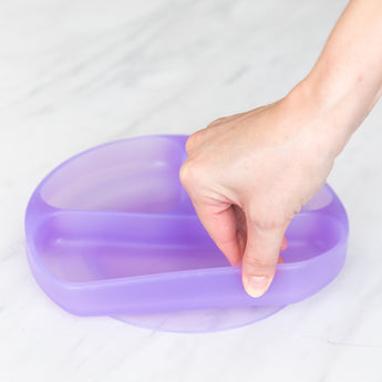 A hand holds a Bumkins Silicone Grip Dish: Purple Jelly, demonstrating the suction cup bottom on a marble surface.