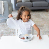 A smiling girl in a white shirt holds the lid of a Bumkins Silicone Grip Dish with Lid (3 Section): Marble at a table in a living room.