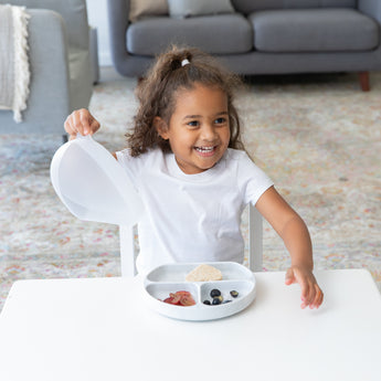 A smiling girl in a white shirt holds the lid of a Bumkins Silicone Grip Dish with Lid (3 Section): Marble at a table in a living room.