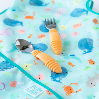 Close-up of Bumkins Spoon + Fork: Tangerine on top of a whale and ocean themed bib on a white background.