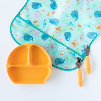 Bumkins Silicone Grip Dish: Tangerine rests on a white background among a whale print bib and orange utensils.