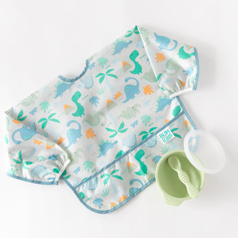 A bib from Bumkins SuperBib® and Sleeved Bib 3-Piece Set: Hangry, Dinosaurs, Blue Tropics lays next to a green bowl & spoon on white.