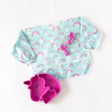 Bumkins Silicone Grip Dish Special Edition: Unicorn sits next to a blue rainbow patterned bib & pink toddler utensils on a white background.