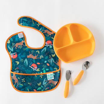 A Bumkins SuperBib® 3 Pack: All Together Now lays flat on a white background next to an orange divided plate and orange utensils.