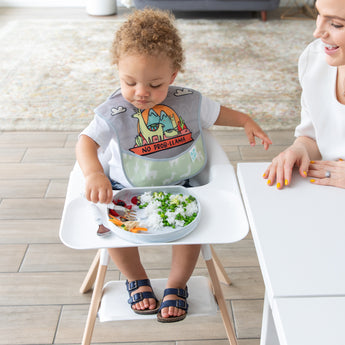 A boy wearing a llama bib in a high chair eats rice & peas from a Bumkins Silicone Grip Plate: Marble as a smiling woman looks on.