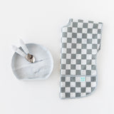 Toddler utensils rest inside Bumkins Silicone Grip Plate: Marble next to a folded gray & white checkered bib on a white background.