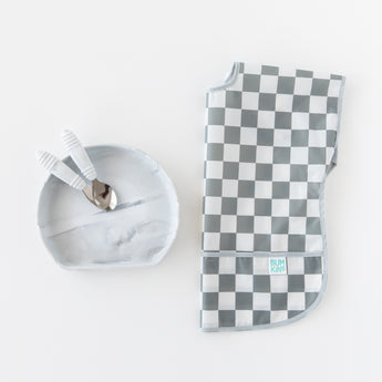 Toddler utensils rest inside Bumkins Silicone Grip Plate: Marble next to a folded gray & white checkered bib on a white background.