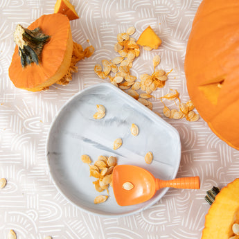 Bumkins Silicone Grip Plate: Marble sits among pumpkin pieces and seeds on a beige patterned mat with an orange scoop.