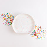 Bumkins Silicone Grip Plate: Vanilla Sprinkle lays on top of a pile of sprinkles in a diagonal line on a white background.