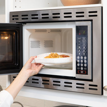 A hand placing a Bumkins Silicone Grip Plate: Vanilla Sprinkle with pasta into an open microwave.
