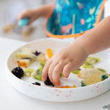 Close-up of a child reaching for fruit from a Bumkins Silicone Grip Plate: Vanilla Sprinkle while wearing a colorful bib.