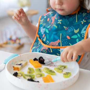 A child in a colorful bib enjoys fruit, cake, and berries from a Bumkins Silicone Grip Plate: Vanilla Sprinkle with a blue spoon.