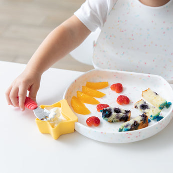 A child in a speckled bib enjoys cake from a Bumkins Silicone Grip Plate: Vanilla Sprinkle, using a red spoon for yogurt in a yellow cup.