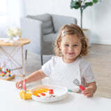 A toddler beams while sitting at a white table, holding a fork and enjoying fruit from a Bumkins Silicone Grip Plate: Vanilla Sprinkle.